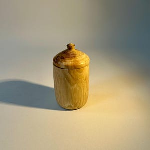 Może przedstawiać: Ręcznie wykonany drewniany pojemnik z pokrywką. Cylindryczny słoik wykonany jest z jasnego drewna, z widocznymi wzorami słojów. Pokrywka ma zaokrągloną górę i dobrze pasuje do słoika. Pojemnik jest prawdopodobnie używany do przechowywania.