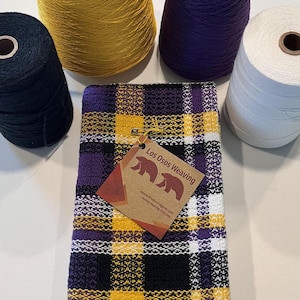 May include: A folded, woven plaid towel in yellow, purple, black, and white, with a tag that reads "Los Osos Weaving." Spools of yellow, purple, black, and white thread are arranged around the towel. The towel's pattern is a grid of squares.