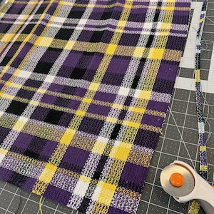 May include: A square piece of woven fabric featuring a plaid pattern in shades of purple, yellow, black, and white. A rotary cutter with an orange blade is positioned near the edge of the fabric, which rests on a dark gray cutting mat.