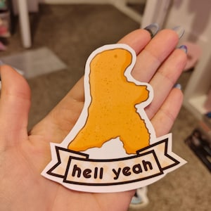 Peut inclure: Un autocollant représentant un nugget de poulet en forme de dinosaure de dessin animé avec le texte "hell yeah" sur une bannière.