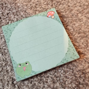 Może przedstawiać: Zielony notes z żabą trzymającą serce i czerwonym grzybem w prawym górnym rogu. Notes ma liniowany papier i zieloną ramkę z kwiatowym wzorem.