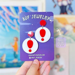 May include: A pair of circular white earrings featuring a bright red exclamation point design. The earrings are attached to a purple display card labeled KGF JEWELRY with the text KEYCHAIN AND PIN OPTIONS AVAILABLE AS WELL! at the side.