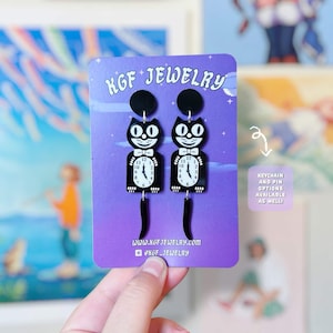 May include: A pair of black and white cat-shaped dangle earrings featuring a clock face on the cat's body and a dangling tail. The earrings are displayed on a purple KGF Jewelry card with the text Keychain and pin options available as well.