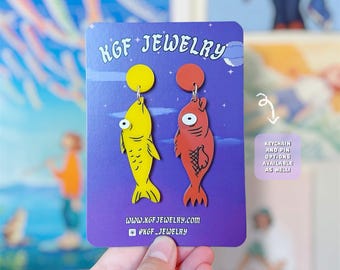 Puede incluir: Un par de pendientes colgantes con forma de pez sobre una tarjeta de presentación morada con la etiqueta KGF JEWELRY. Un pez es amarillo brillante y el otro es rojo anaranjado, ambos con grandes ojos blancos. Incluye la URL del sitio web.