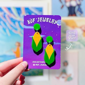 Pode incluir: Um par de brincos pendentes em forma de milho num cartão de apresentação roxo da KGF Jewelry. Os brincos apresentam uma espiga de milho amarela com folhas verdes e um pequeno pino circular verde. O cartão inclui o texto KGF JEWELRY e redes sociais.