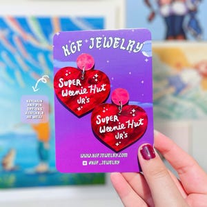 Può includere: Due orecchini in acrilico rosso a forma di cuore fissati su un cartoncino viola. Gli orecchini presentano la scritta bianca Super Weenie Hut Jr's. Il cartoncino riporta il testo KGF Jewelry, il sito www.kgfjewelry.com e @kgf_jewelry.