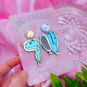 May include: A pair of blue and silver earrings featuring a melting clock design. The earrings are made of wood and have a glossy finish. The earrings are displayed on a white card with the text "KGF JEWELRY" and a website address.