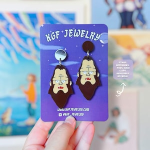 Könnte beinhalten: Ein Paar braune Ohrringe in Bigfoot-Form auf einer violetten KGF Jewelry-Präsentationskarte. Die Ohrringe zeigen eine Cartoon-Figur mit Bart und zwei Zähnen. Die Karte enthält den Text KGF JEWELRY, die Website und einen Hinweis auf weitere Artikel.