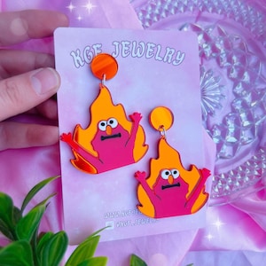 Könnte beinhalten: Ein Paar ausgefallene Ohrringe mit einem Cartoon-Charakter im Flammen-Design. Die Ohrringe sind rosa, orange und rot, mit einem kreisförmigen orangefarbenen Stecker. Auf einer rosa Karte steht der Text "KGF JEWELRY".
