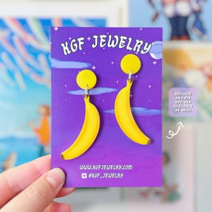 以下が含まれることがあります： KGF JEWELRYという文字が記された紫色の台紙に固定された、黄色のバナナ型イヤリング。台紙には星空のデザインが施されており、ウェブサイトのURLと、キーホルダーやピンのオプションも利用可能である旨が記載された小さなボックスが表示されています。
