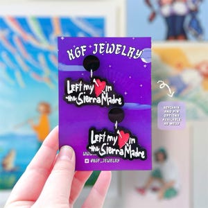 May include: A pair of black acrylic earrings on a purple display card. Each earring features a red heart pierced by an arrow and the white text "Left my in the Sierra Madre". The card displays "KGF JEWELRY" at the top and social media info.