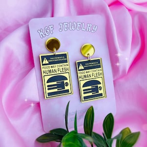 May include: A pair of gold earrings with black warning signs that read "This premises is under investigation. Food may contain human flesh." The signs have a stylized image of a hamburger.