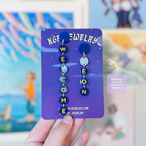 May include: A pair of mismatched dangle earrings on a purple display card. The left earring features seven blue circular charms spelling WELCOME in yellow letters. The right earring has four blue circular charms spelling LEON in yellow letters.