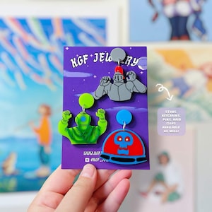 May include: Three acrylic earrings on a purple display card labelled KGF JEWELRY. The earrings feature a grey knight, a bright green diver, and a red and blue skull in a dome. A small text box on the right reads: STUDS, KEYCHAINS, PINS, HAIR CLIPS AVAILABLE AS WELL!