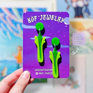 May include: A pair of bright green celery stalk earrings attached to a purple display card with the text KGF JEWELRY. The card includes a small box with an arrow pointing to the text KEYCHAIN AND PIN OPTIONS AVAILABLE AS WELL.