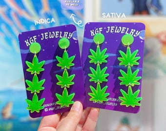 May include: Two pairs of bright green acrylic cannabis leaf earrings displayed on purple KGF Jewelry backing cards. The left card is labeled Indica and the right card is labeled Sativa. Each earring features three stacked leaf charms hanging from a circular stud.