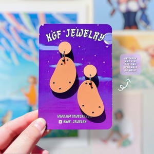 Peut inclure: Une paire de boucles d'oreilles en forme de pomme de terre couleur pêche sur une carte de présentation violette marquée KGF JEWELRY. Les boucles comportent un élément circulaire supérieur relié à une base ovale irrégulière avec des points sombres. La carte affiche WWW.KGFJEWELRY.COM et @KGF_JEWELRY.