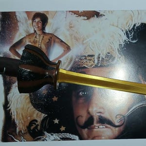 Puede incluir: Un abrecartas decorativo con una hoja dorada y un mango marrón oscuro. El mango tiene un diseño único con una firma. El fondo presenta un cartel de película con imágenes de actores.