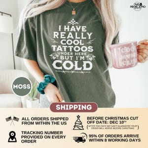 May include: A moss green short-sleeve t-shirt featuring white text that reads I HAVE REALLY COOL TATTOOS UNDER HERE BUT I'M COLD, accented with small floral illustrations and a barbed wire graphic. The shirt is paired with a teal scrunchie.