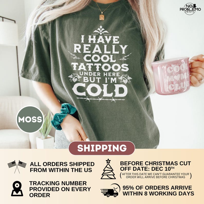 May include: A moss green short-sleeve t-shirt featuring white text that reads I HAVE REALLY COOL TATTOOS UNDER HERE BUT I'M COLD, accented with small floral illustrations and a barbed wire graphic. The shirt is paired with a teal scrunchie.