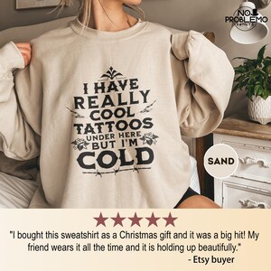 May include: A sand-colored crewneck sweatshirt featuring black text that reads: I HAVE REALLY COOL TATTOOS UNDER HERE BUT I'M COLD. The design includes decorative floral accents and a barbed wire graphic at the bottom of the lettering.