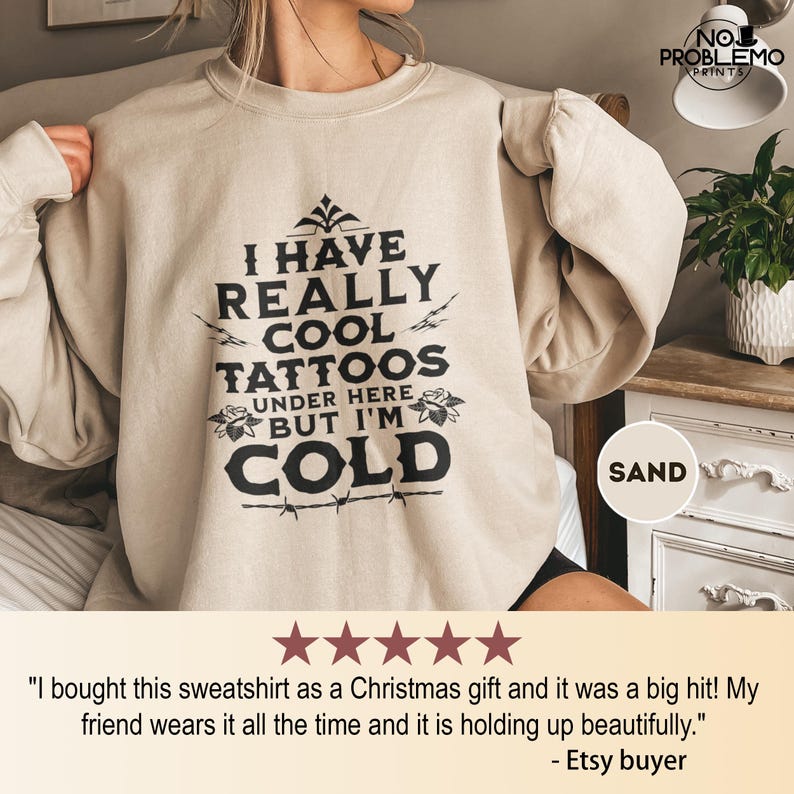 May include: A sand-colored crewneck sweatshirt featuring black text that reads: I HAVE REALLY COOL TATTOOS UNDER HERE BUT I'M COLD. The design includes decorative floral accents and a barbed wire graphic at the bottom of the lettering.