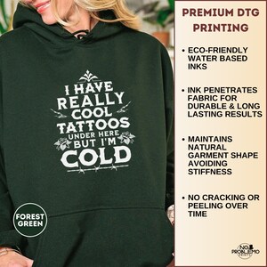 May include: A forest green hooded sweatshirt featuring white text that reads I HAVE REALLY COOL TATTOOS UNDER HERE BUT I'M COLD, accented with small rose and barbed wire graphics. The side panel lists premium DTG printing features.