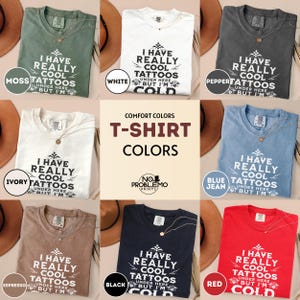 May include: A collection of eight short-sleeved t-shirts in moss, white, pepper, ivory, blue jean, espresso, black, and red. Each shirt features the text I HAVE REALLY COOL TATTOOS UNDER HERE BUT I'M COLD printed in a bold, dark font on the front.