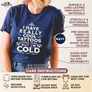 May include: A navy blue short-sleeved t-shirt featuring white text that reads I HAVE REALLY COOL TATTOOS UNDER HERE BUT I'M COLD. The shirt includes graphic details of roses and barbed wire. The image displays product features and care instructions.