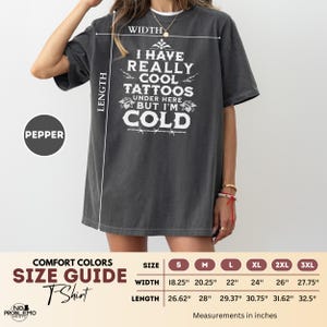 May include: A charcoal grey short-sleeve t-shirt featuring white text that reads I HAVE REALLY COOL TATTOOS UNDER HERE BUT I'M COLD. A size guide chart is included, showing width and length measurements in inches for sizes S through 3XL.