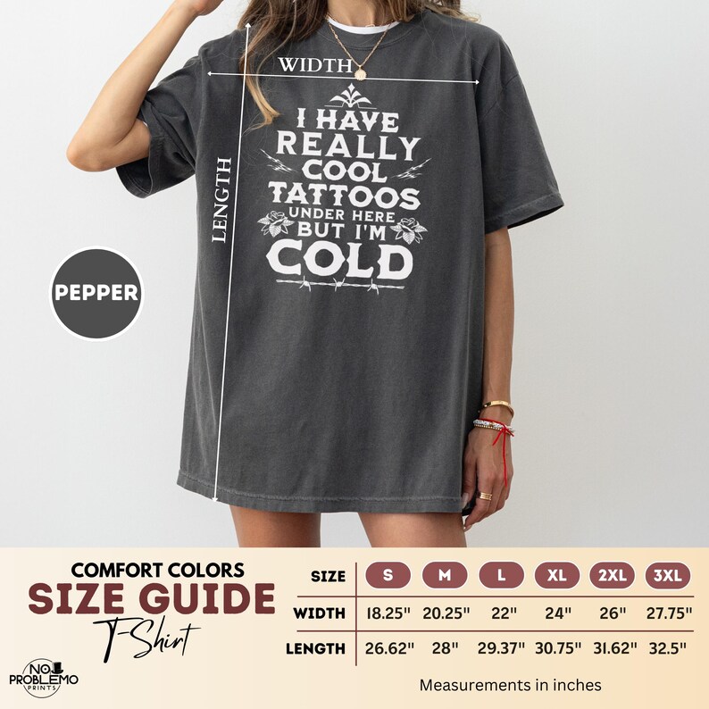 May include: A charcoal grey short-sleeve t-shirt featuring white text that reads I HAVE REALLY COOL TATTOOS UNDER HERE BUT I'M COLD. A size guide chart is included, showing width and length measurements in inches for sizes S through 3XL.