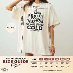 May include: A natural-colored t-shirt featuring black text that reads I HAVE REALLY COOL TATTOOS UNDER HERE BUT I'M COLD, accompanied by a size guide chart with width and length measurements in inches for sizes S through 3XL, Bella+Canvas 3001.
