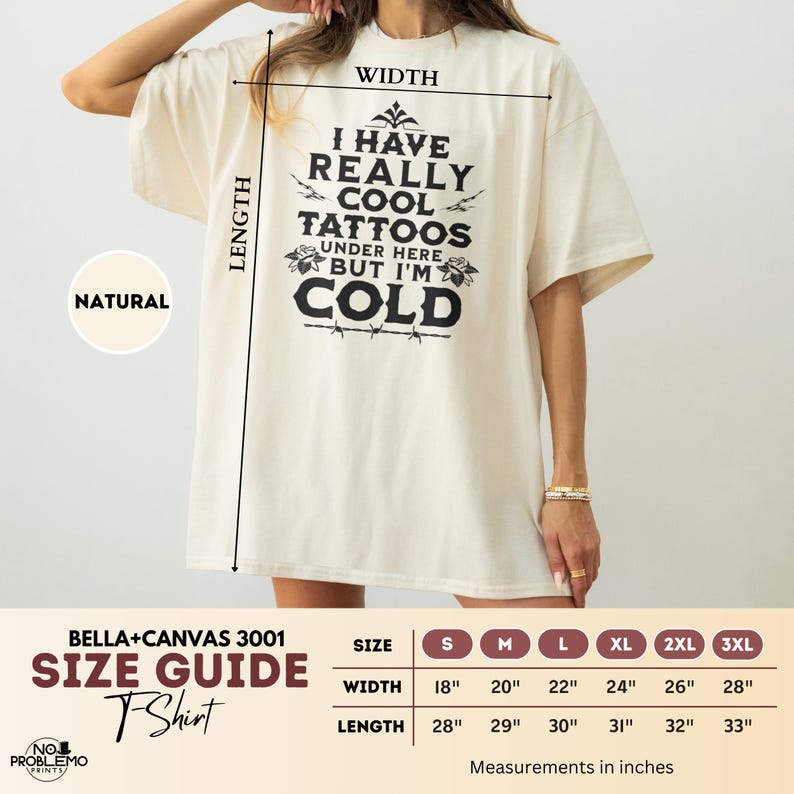 May include: A natural-colored t-shirt featuring black text that reads I HAVE REALLY COOL TATTOOS UNDER HERE BUT I'M COLD, accompanied by a size guide chart with width and length measurements in inches for sizes S through 3XL, Bella+Canvas 3001.