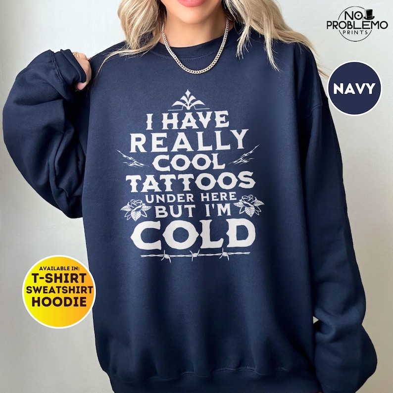 May include: A navy blue crewneck sweatshirt featuring white text that reads I HAVE REALLY COOL TATTOOS UNDER HERE BUT I'M COLD, accented with small floral illustrations and a barbed wire graphic below the final word, worn with a silver chain necklace.