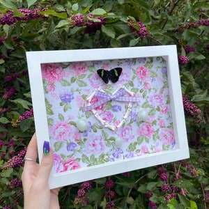 May include: A white framed artwork featuring a floral patterned background with pink and purple flowers. A black butterfly, a purple and white ribbon, and white stones are arranged on the artwork. The artwork is held in front of a bush with purple berries.