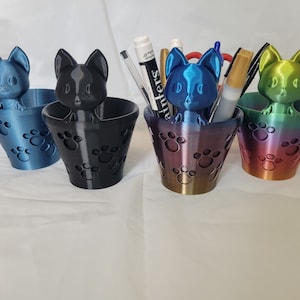 May include: Four colorful pen holders, each featuring a cat-shaped top. The holders are in shades of blue, black, iridescent purple, and rainbow. Paw prints decorate the sides. Some holders contain pens and markers.