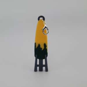 May include: A yellow and green corn on the cob keychain with a silver metal clasp. The corn is standing on a grey stand.