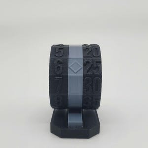 May include: A black and gray 3D-printed die with numbers from 5 to 35. The die is cylindrical with a gray band and sits on a black base. Numbers are printed in white.