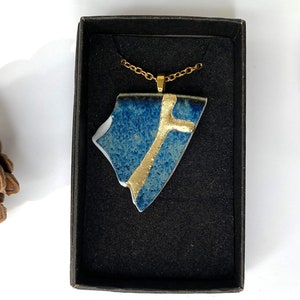 May include: A blue and gold ceramic pendant necklace in a black box. The pendant is a shard of pottery with gold accents. The necklace is on a gold chain.