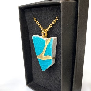 May include: A turquoise and gold pendant necklace displayed in a black gift box. The pendant is a unique, irregularly shaped piece with a gold-colored design. The necklace has a delicate gold chain. The background is white with a pine cone.