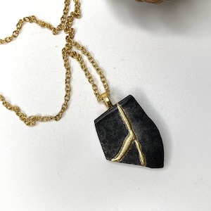 Könnte beinhalten: Eine goldene Kettenkette mit einem schwarzen Anhänger mit goldenem Design. Der Anhänger hat die Form eines Blattes.
