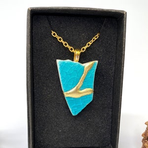 May include: A turquoise blue ceramic pendant with a gold accent, hanging from a gold chain in a black box. The pendant is shaped like a shard of glass.