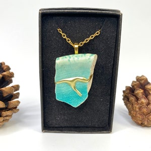 May include: A turquoise ceramic pendant with a gold accent, hanging from a gold chain in a black box. The pendant has a crackled glaze and a unique shape.