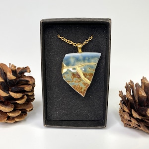 May include: A gold chain necklace with a blue and brown ceramic pendant in a black box. The pendant has a crackled glaze and gold accents.