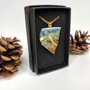 Könnte beinhalten: Eine goldene Kettenkette mit einem blauen und braunen Keramik-Anhänger in einer schwarzen Schachtel. Der Anhänger hat eine einzigartige, unregelmäßige Form und goldene Akzente.