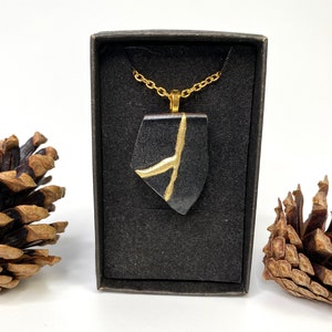 May include: A black pendant necklace with a gold chain and a geometric design. The pendant is made of black resin with a gold accent. The necklace is displayed in a black box.