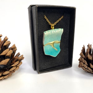 May include: A turquoise and white ceramic pendant necklace with a gold-colored chain. The pendant is a broken piece of ceramic with a gold accent. The necklace is displayed in a black box, with pine cones on either side.