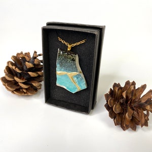 May include: A gold chain necklace with a blue and black ceramic pendant in a black box. The pendant is a broken piece of pottery with gold accents.
