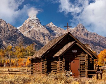 May include: A wooden log cabin church with a cross on the roof, set against a backdrop of snow-capped mountains and fall foliage. The sign on the front of the church reads "Chapel of the Transfiguration".