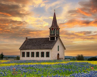 May include: A white stone church with a tall steeple and a brown roof stands in a field of bluebonnet wildflowers. The sky is a vibrant orange and pink, creating a beautiful sunset backdrop.